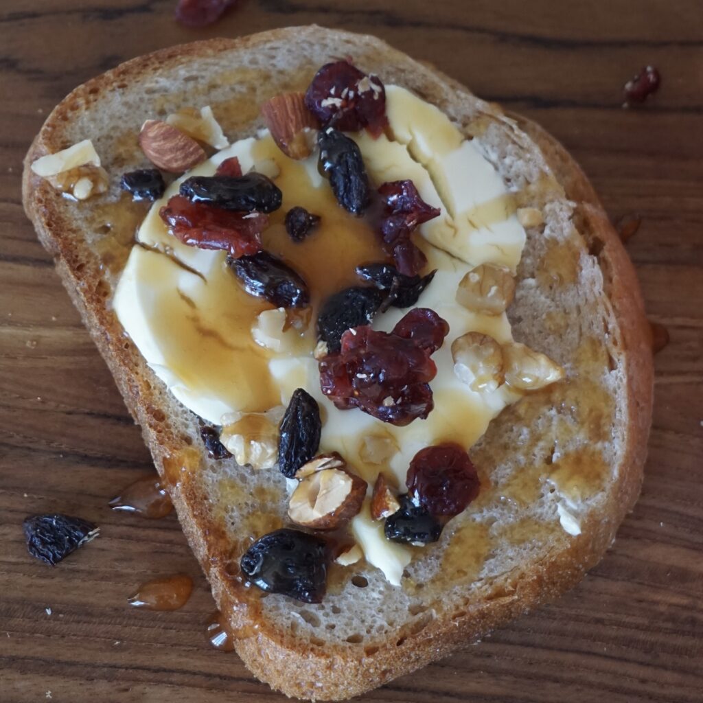 This is a recipe for toast made with rye bread made in a home bakery, topped with cream cheese, dried fruit, and nuts, and drizzled with plenty of Manuka honey.